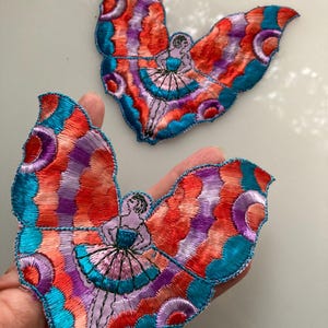 May include: Two embroidered patches featuring a figure with large, colorful butterfly wings. The wings display a vibrant pattern of red, orange, purple, and teal thread. The central figure wears a dress and has dark hair, with detailed stitching.
