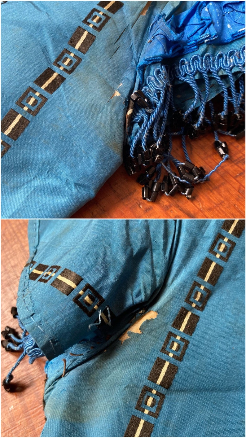 May include: A close-up of a blue fabric with a black and white geometric pattern. The fabric has a frayed edge and is decorated with black beads.