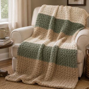 Puede incluir: Una manta de punto grueso con rayas horizontales en crema, verde salvia y beige. La manta presenta un patrón texturizado de tejido abierto y está colocada sobre un sillón acolchado, destacando su construcción de hilo grueso y tonos neutros.