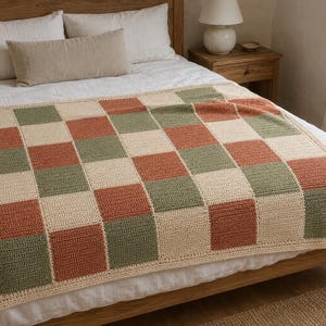 May include: A rectangular crochet blanket featuring a repeating checkerboard pattern of cream, sage green, and terracotta orange squares. The blanket has a solid cream-colored border and is constructed with a visible textured yarn stitch pattern.