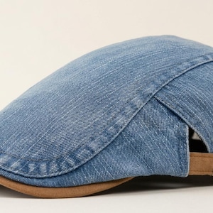 Op de afbeelding: Een lichtblauwe denim baret met een gebogen klep en een bruine, suède-achtige rand. De achterkant van de pet is voorzien van een verstelbare bruine riem met een metalen gesp voor een goede pasvorm. De stof heeft een klassieke denim textuur.
