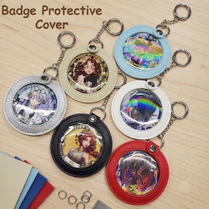 Puede incluir: Seis fundas protectoras circulares para insignias con llaveros de metal en plata, crema, negro, azul claro, blanco y rojo. Cada funda tiene una ventana transparente con una ilustración. El texto Badge Protective Cover es visible.