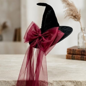 May include: A black velvet witch hat featuring a large, sheer burgundy organza bow attached to the front. The bow has long, flowing ribbons that drape downward from the brim of the hat, creating a layered and textured appearance on the accessory.