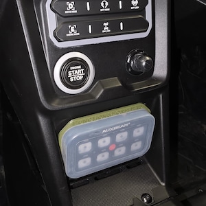 May include: A black vehicle center console featuring a silver-rimmed engine start-stop button, a control panel with drive mode and differential lock icons, and a rectangular Auxbeam switch panel with eight buttons mounted in a custom green bracket.