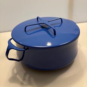 Puede incluir: Una olla redonda de metal esmaltado en color azul cobalto brillante con dos asas laterales y una tapa a juego. La tapa presenta un distintivo mango en forma de cruz centrado en la parte superior. La olla tiene un acabado liso y uniforme.