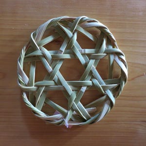 以下が含まれることがあります： 淡い緑色とベージュ色の植物繊維を編み込んで作られた円形の鍋敷き。幾何学的な模様が特徴で、編み込まれた円形の枠の中に、6つの角を持つ星型のデザインが中央に配置されています。