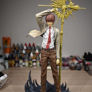 May include: A standing anime-style figure of a person with brown hair, wearing a white shirt, red tie, and beige blazer. The figure holds a large, golden, star-shaped staff above their head and stands on a dark, textured, swirling base.