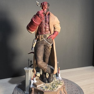May include: A Hellboy figurine featuring a red-skinned character with a large stone hand, wearing a tan trench coat and dark trousers. The figure stands on a detailed base with a skull, a book, and candles, holding a lit cigar to its mouth.