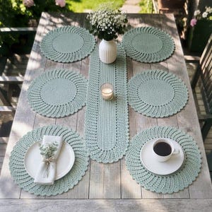 May include: A set of six round, sage green crocheted placemats and a matching rectangular table runner with scalloped edges. The runner features a central vertical pattern, while the placemats display a radiating, spiral-like stitch design.
