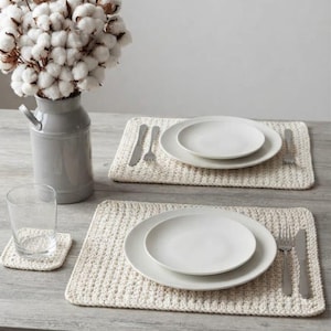 May include: Two rectangular cream-coloured crocheted placemats and one matching square coaster. Each placemat holds a large white dinner plate topped with a smaller white salad plate, a silver fork, and a silver knife. A grey pitcher holds white cotton stems.