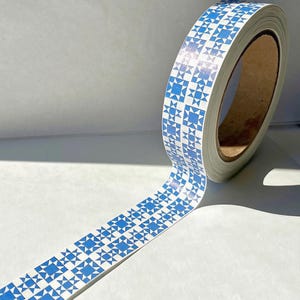 May include: A roll of adhesive tape featuring a repeating blue and white geometric quilt block pattern. The tape has a smooth, glossy finish and is wound around a central cardboard core, with a portion of the tape extended outward.