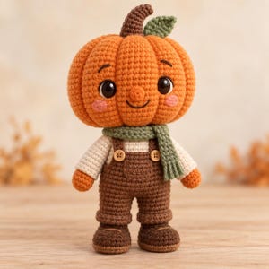 Puede incluir: Una muñeca de ganchillo con una cabeza de calabaza naranja grande con una pequeña hoja verde, ojos negros y mejillas rosadas. La muñeca viste una camisa de manga larga color crema, un peto marrón con dos botones de madera, una bufanda verde y zapatos.