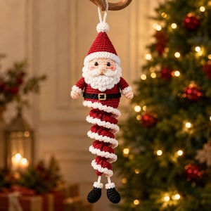 Puede incluir: Un adorno de Papá Noel tejido a ganchillo con gorro rojo y blanco, barba blanca, chaqueta roja con cinturón negro y hebilla dorada, y un cuerpo largo en espiral de color rojo y blanco que termina en dos pequeñas botas negras.