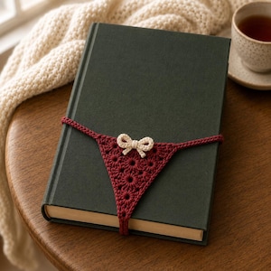 May include: A dark green hardcover book wrapped with a dark red crocheted band featuring a small cream-colored bow at the center. The band is constructed with a repeating floral-like stitch pattern and wraps horizontally around the book cover.