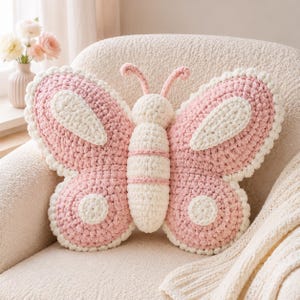 May include: A butterfly-shaped crochet pillow featuring a central body in cream and pink, with large wings in soft pink and cream accents. The pillow has two small antennae on top and a textured, chunky yarn construction throughout the design.