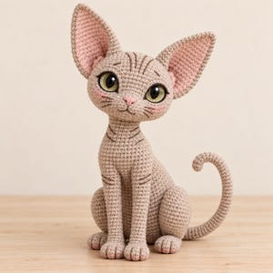 May include: A crocheted amigurumi figure of a hairless Sphynx cat in a light beige yarn. The cat features large pointed ears with pink inner detailing, oversized green eyes, a small pink nose, and embroidered facial wrinkles and whiskers.