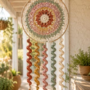 May include: A circular wall hanging featuring a central crochet mandala in shades of cream, rust, yellow, pink, and sage green. Five long, spiral-shaped crochet strands hang from the bottom, each ending with a wooden bead and a matching yarn tassel.