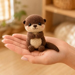 Könnte beinhalten: Ein kleines, handgefertigtes Häkel-Otter-Spielzeug aus dunkelbraunem Garn für Kopf, Rücken und Schwanz sowie beigefarbenem Garn für Gesicht und Bauch. Der Otter hat zwei schwarze Kunststoffaugen, eine kleine gestickte Nase und rosa Wangen.