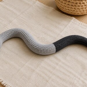 May include: A crocheted snake toy featuring a light gray head and body that transitions into a dark charcoal gray tail. The snake has black bead eyes, a small pink stitched mouth, and a protruding pink forked tongue made of yarn.