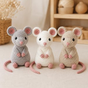 Peut inclure: Trois figurines de souris crochetées côte à côte. La souris de gauche est grise, celle du centre est blanche et celle de droite est beige. Chaque souris présente des oreilles rondes bordées de rose, des yeux en perles noires et une queue rose.