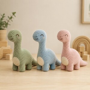 Peut inclure: Trois dinosaures en crochet de couleur vert sauge, bleu clair et rose pâle. Chaque dinosaure présente un long cou, un corps arrondi avec une pièce circulaire de couleur crème sur le côté, et des détails faciaux brodés incluant les yeux et un sourire.