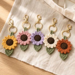 May include: Five crochet flower keychains with gold-tone metal lobster clasps and rings. Each keychain features a dark brown center, petals in yellow, pink, light purple, cream, or rust orange, and a small green leaf attached at the bottom.
