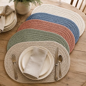 Op de afbeelding: Een set van vijf ovale, gehaakte placemats in crème, blauw, terracotta, saliegroen en beige. De placemats hebben een dicht, gestructureerd steekpatroon. Eén beige placemat is gedekt met een wit keramieken bord, een linnen servet, een vork en een lepel.