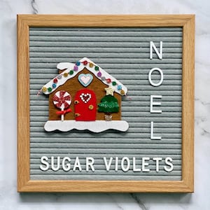 May include: A gray letter board with a felt gingerbread house applique. The house features red and white details, candy, and a small Christmas tree.  The words 
