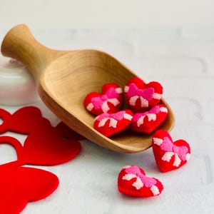 May include: A wooden scoop filled with red heart-shaped ornaments with pink and white embroidered details. Several hearts are scattered around the scoop. The background is a white brick wall.