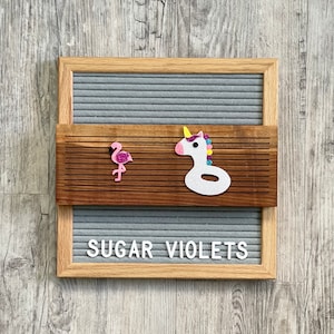 May include: A gray letter board with a wooden frame and a brown wood strip across the middle. The board has white letters spelling out "SUGAR VIOLETS". There is a pink flamingo and a white unicorn float on the wood strip.