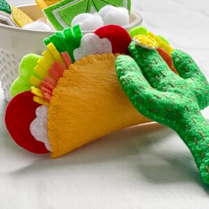 Op de afbeelding: Een vilten taco met kleurrijke toppings, waaronder rode, witte, groene en gele elementen. Een groene vilten cactus staat naast de taco. De taco is geel en heeft een gestikt ontwerp.