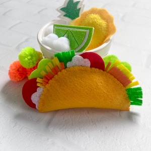 Op de afbeelding: Een vilten taco met kleurrijke toppings, waaronder rode tomaten, groene sla en witte wolken. Een kleine kom bevat een limoenwig, een gele bloem en een ananas. Oranje en groene pompons staan links.