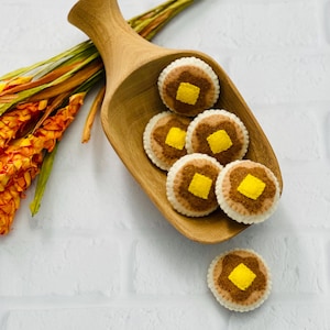 May include: A wooden scoop filled with felt pancakes topped with yellow butter squares. The pancakes have a brown center and a cream-colored edge. A sprig of orange and yellow flowers and a long, thin, yellow leaf are in the background. The background is a white brick wall.