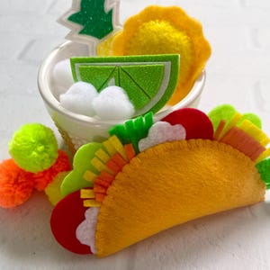 Op de afbeelding: Een vilten taco met kleurrijke vullingen, naast een kleine kom met vilt ananas, limoen en witte pompons. Heldere gele, oranje, groene en rode viltstukken cre&euml;ren de ingredi&euml;nten van de taco. Een paar neon groene en oranje pompons liggen in de buurt.