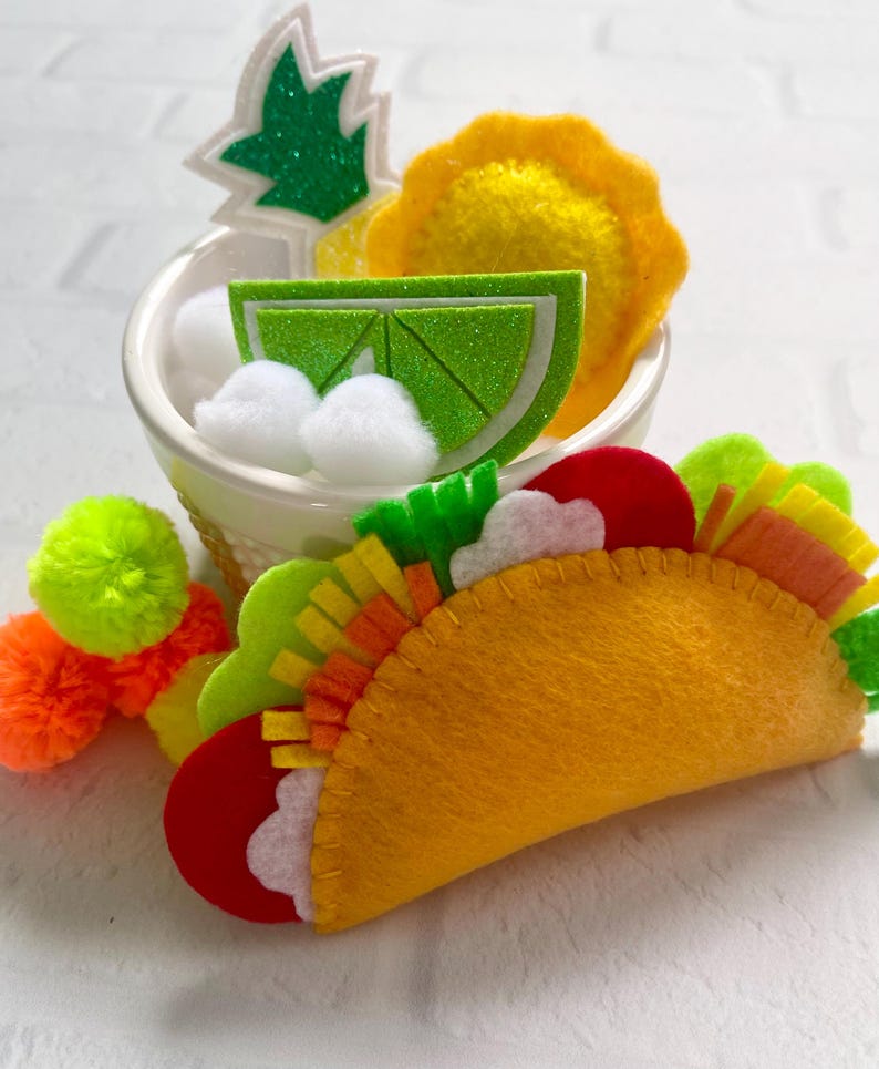 Op de afbeelding: Een vilten taco met kleurrijke vullingen, naast een kleine kom met vilt ananas, limoen en witte pompons. Heldere gele, oranje, groene en rode viltstukken cre&euml;ren de ingredi&euml;nten van de taco. Een paar neon groene en oranje pompons liggen in de buurt.