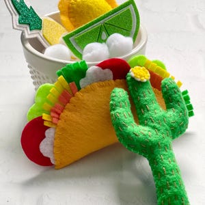Op de afbeelding: Een gele vilten taco met kleurrijke toppings, een groene cactus en een kom met limoen, citroen en andere taco-ingredi&euml;nten. De taco heeft rode, witte, groene en gele toppings. De cactus is groen met gouden stiksels.