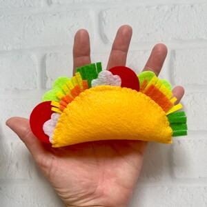 Op de afbeelding: Een vilten taco met gele, rode, groene, oranje en witte componenten. De tacoschelp is geel en de vullingen omvatten rode tomatenschijfjes, witte zure room en groene sla. De taco wordt in een hand gehouden.