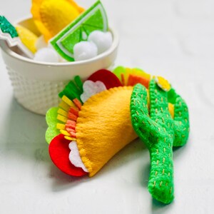 Op de afbeelding: Een vilten taco met gele, rode, witte en groene vullingen, naast een groene vilten cactus. Een witte kom bevat vilten limoenschijfjes, gele items en witte ballen. De items zijn handgemaakt.