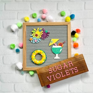 May include: A gray letter board with a yellow and white life preserver, a green cocktail with a cherry and a pink and yellow firework design. A wooden sign with the text "SUGAR VIOLETS" in pink letters.