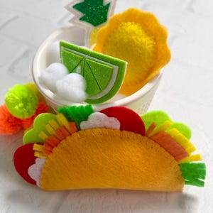 Op de afbeelding: Een vilten taco met kleurrijke toppings, waaronder rode tomaten, witte wolken en groene sla. Een kleine witte kom bevat een limoenschijfje, een ananas en een geel item. De taco is geel en heeft een gestikte rand.