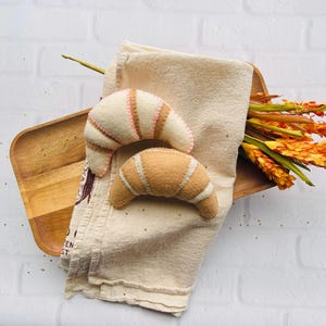 May include: Two crescent-shaped felt croissant toys, one in cream with tan stripes and one in tan with cream stripes, resting on a folded beige cloth napkin atop a wooden serving tray with a cluster of orange and yellow dried flowers.