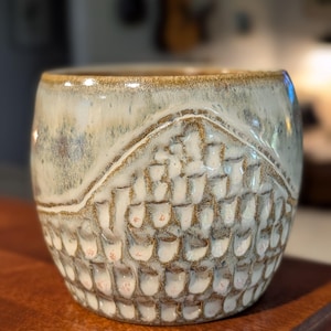 May include: A ceramic mug featuring a two-tone glaze with a smooth, light-colored upper section and a textured, carved lower section. The lower portion displays a repeating, scale-like pattern with brown detailing against a cream-colored base.