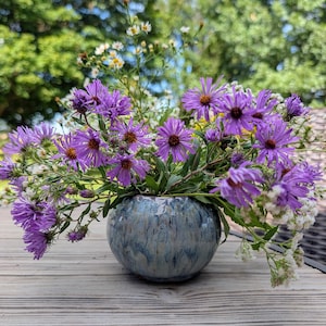 May include: A round, ceramic vase with a glossy, mottled blue and grey glaze finish, filled with a vibrant arrangement of purple aster flowers and small white wildflowers, showcasing a natural, organic aesthetic with varied petal textures.