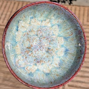 May include: A round ceramic bowl featuring a crystalline glaze with a vibrant blend of turquoise, light blue, and cream colours. The interior surface displays a starburst pattern, while the outer rim is finished with a distinct reddish-brown edge.