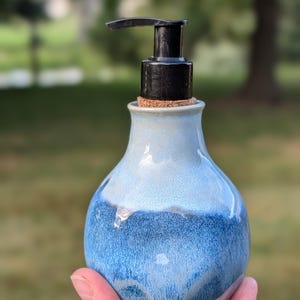 May include: A ceramic soap dispenser with a bulbous base featuring a gradient of light blue at the top transitioning to a deep, speckled navy blue at the bottom. The vessel is topped with a cork stopper and a functional black plastic pump mechanism.