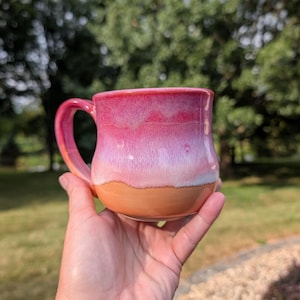 May include: A ceramic mug featuring a gradient glaze that transitions from a deep magenta at the rim to a lighter pink in the center, ending with a solid orange base. The mug has a curved handle on the left side and a smooth, rounded body shape.