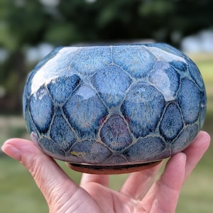 May include: A small, round ceramic bowl featuring a glossy, crystalline glaze in shades of deep blue, light blue, and hints of purple. The surface displays a distinct honeycomb or scale-like pattern with dark outlines, and the base is unglazed terracotta.