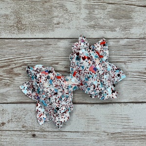 May include: A white, red, and blue glitter bow with a ruffled design. The bow is decorated with small American flag accents.