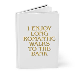 Pode incluir: Um livro de capa dura branco com o texto dourado I ENJOY LONG ROMANTIC WALKS TO THE BANK impresso na capa frontal. O livro está ligeiramente aberto no topo, revelando páginas brancas no interior, e encontra-se em posição vertical.