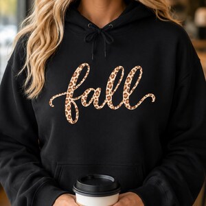 May include: A black hooded sweatshirt featuring the word fall written in a cursive script font with a brown and black leopard print pattern. The hoodie includes a matching black drawstring at the neckline and a front kangaroo pocket.